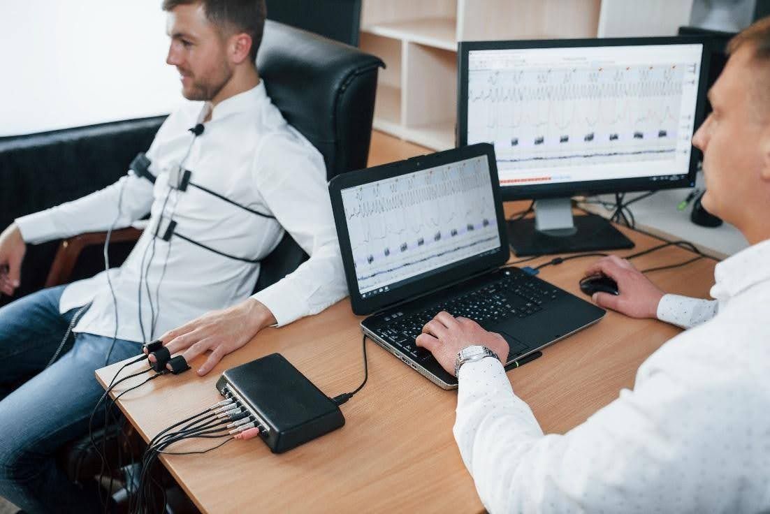 Man connected to polygraph machine; another person operates a laptop displaying charts.