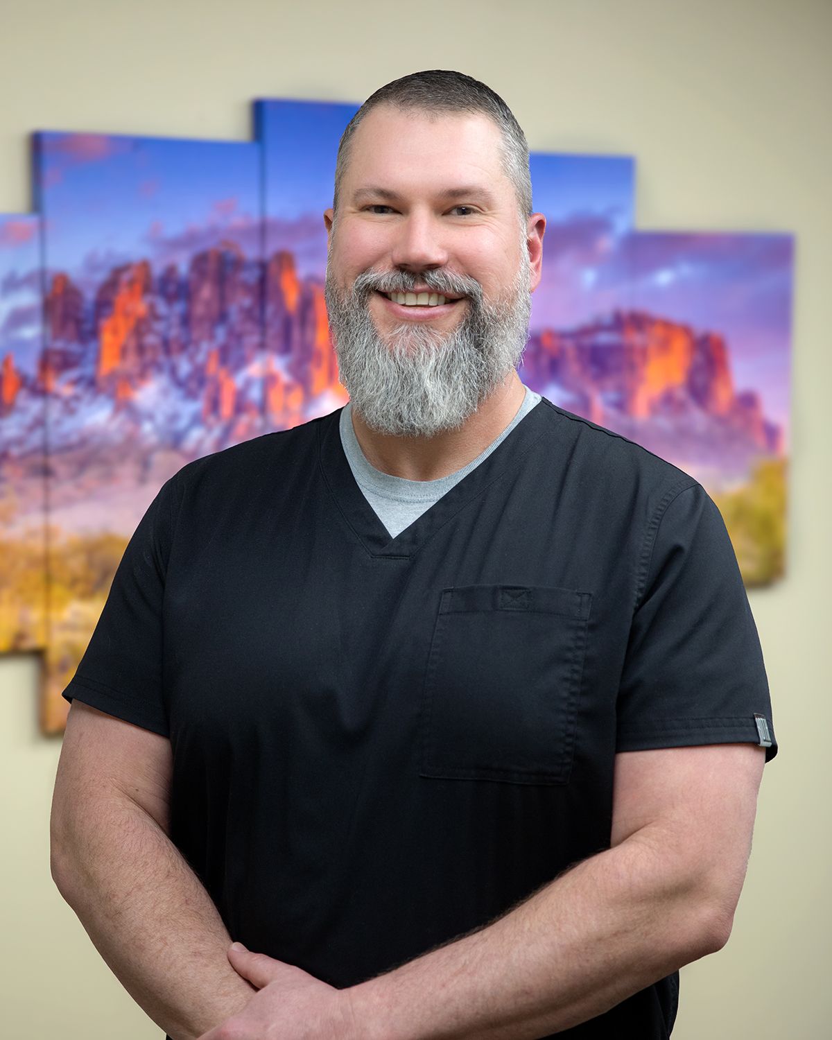 A man with a beard is wearing a black scrub top and smiling.