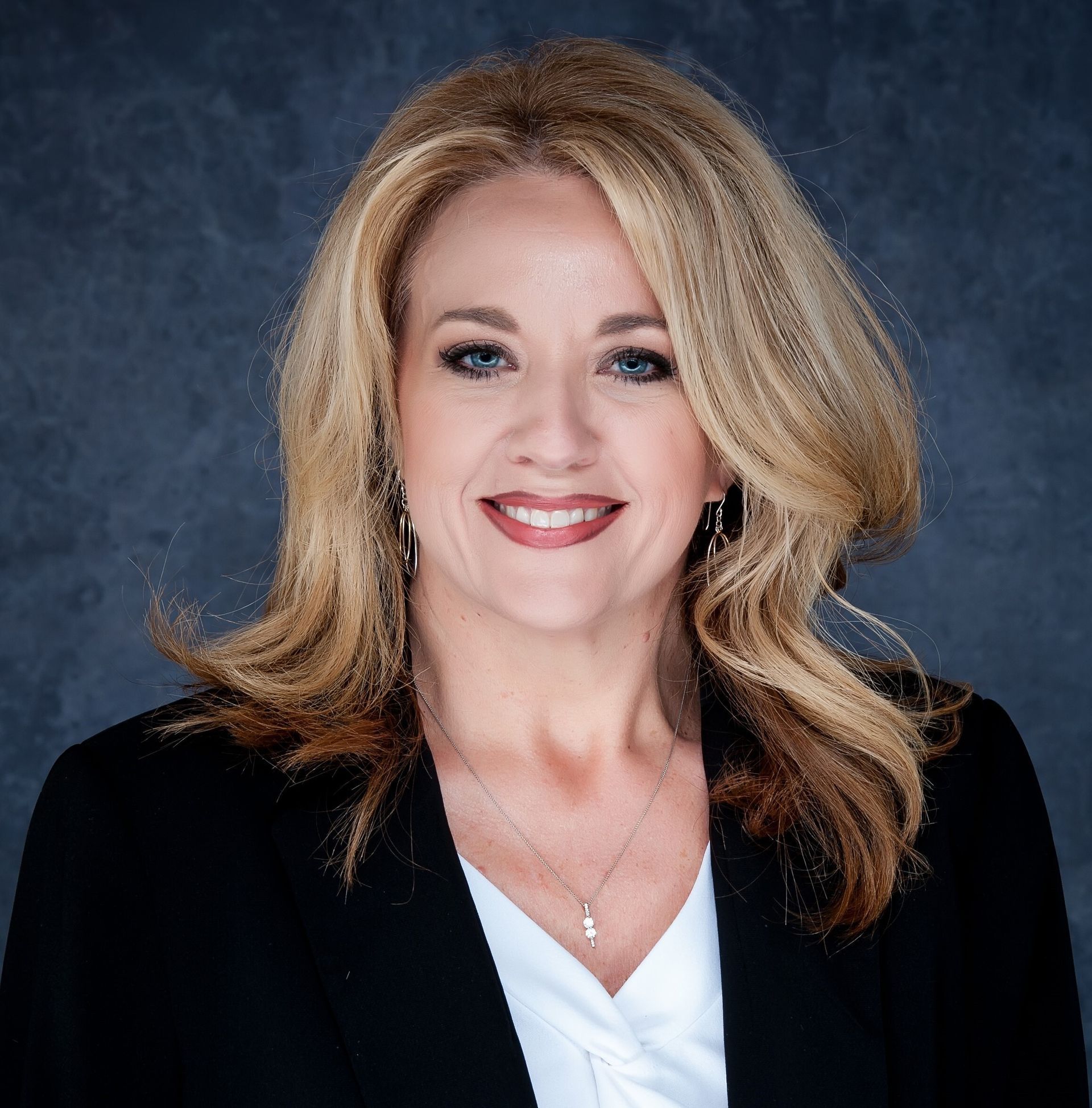 Blonde woman smiling, wearing a black blazer over a white top, against a blurred background.