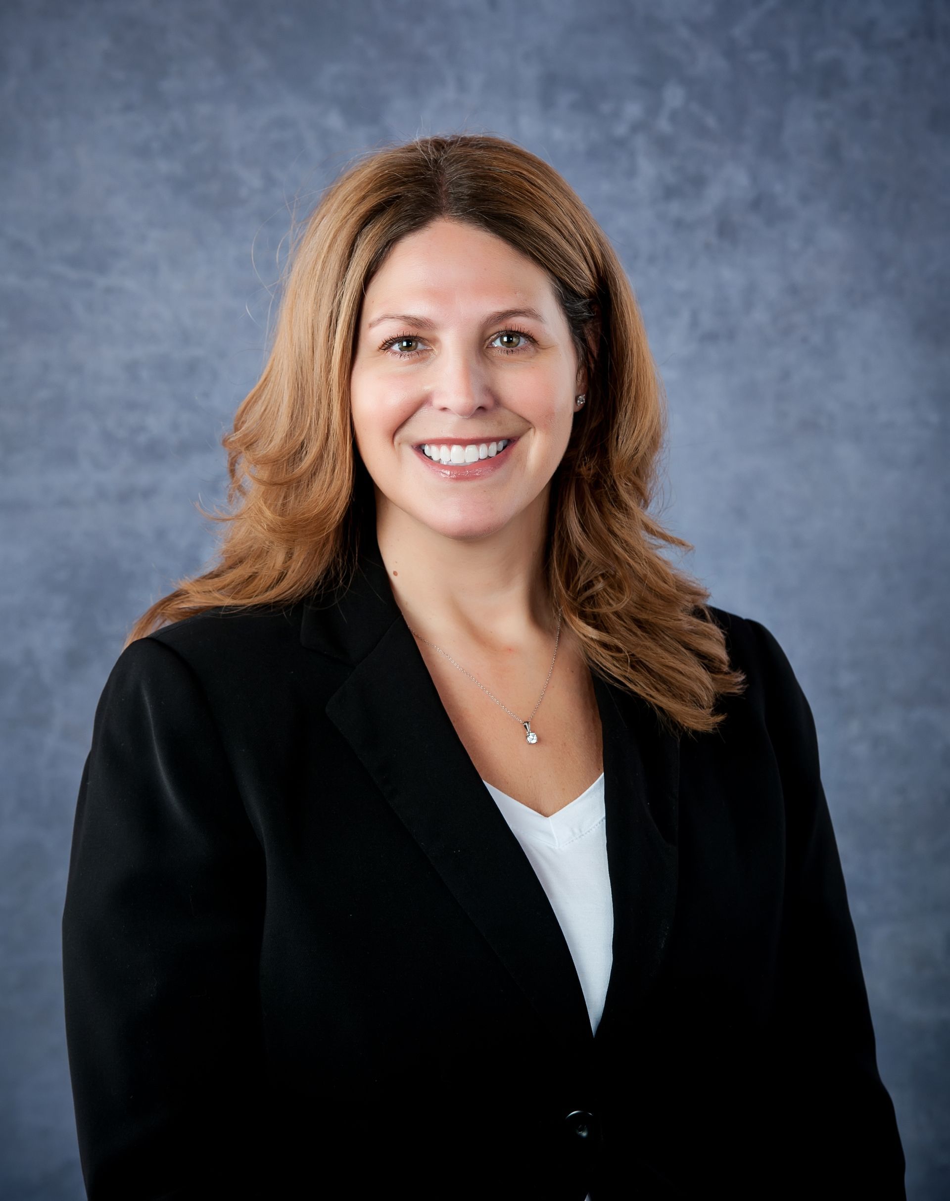 Woman with brown hair in a black suit smiling at the camera, against a blue-gray background.