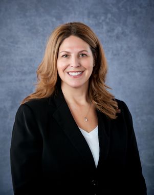 Woman with brown hair in a black suit smiling at the camera, against a blue-gray background.