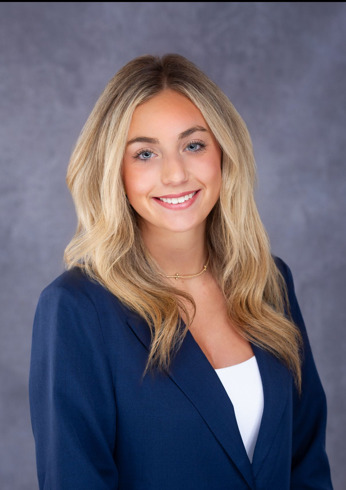 Blonde woman in navy blazer smiles at camera; chest-up shot against gray backdrop.