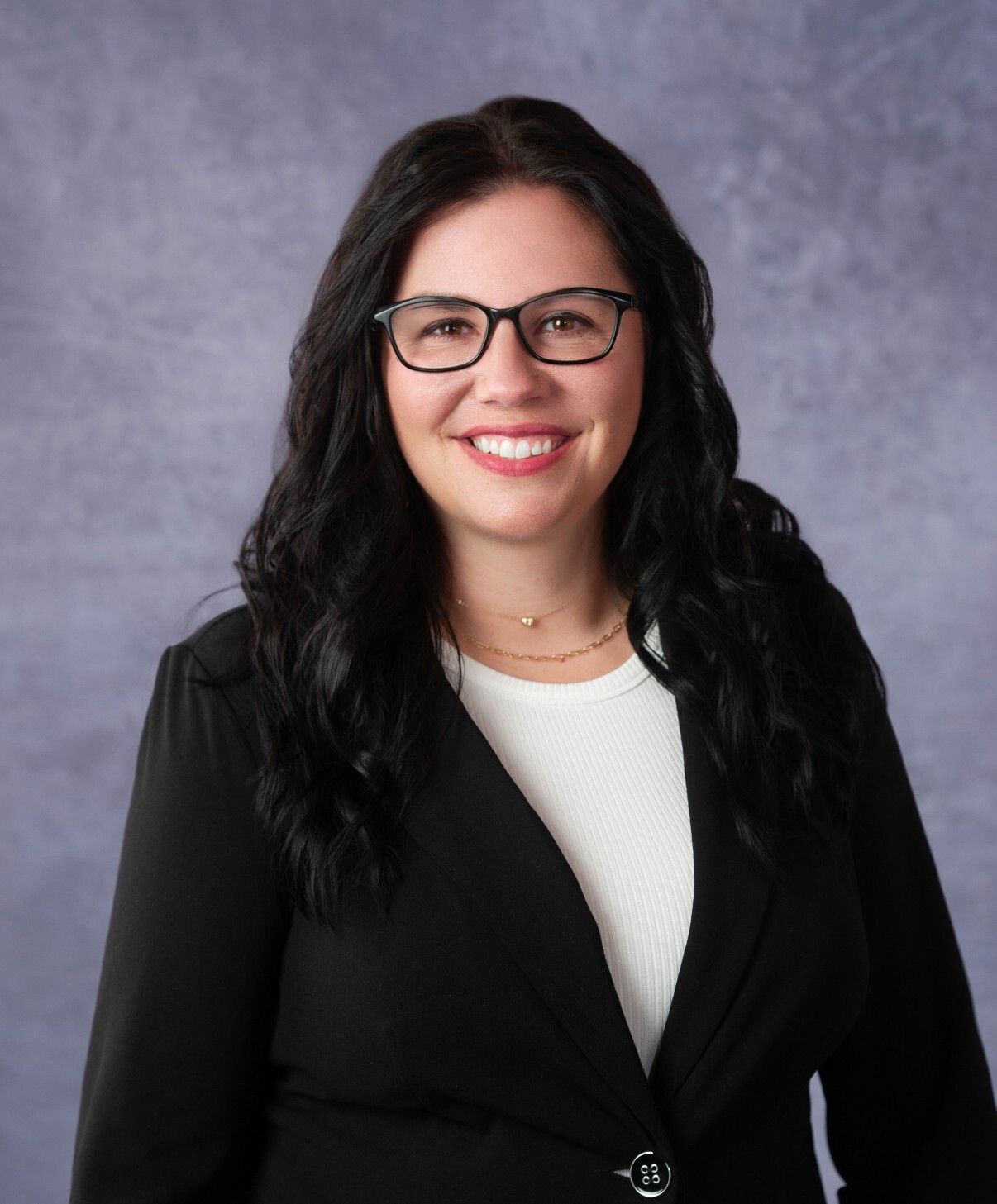 Woman with glasses, dark hair, wearing a black blazer and smiling in front of a blurred blue background.