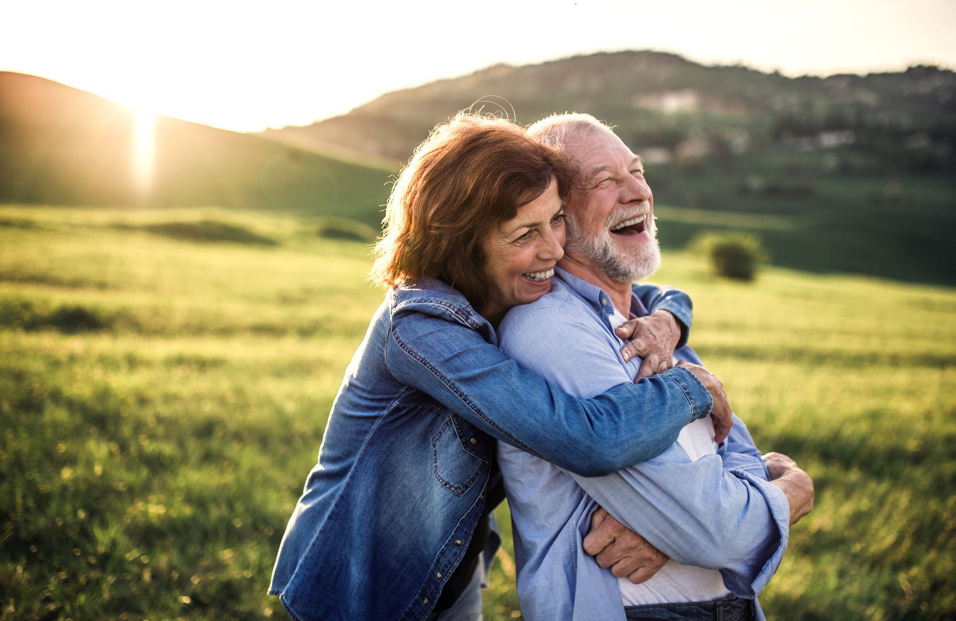 Woman hugging and smiling at an older man, both in a grassy field with a sunset.