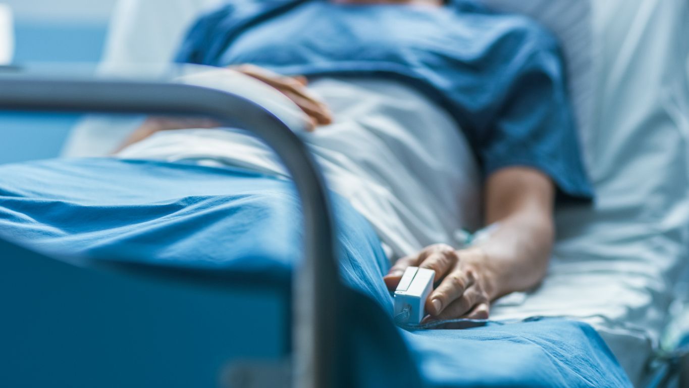 Person in a hospital bed with an IV hand, wearing blue and resting under blue sheets.