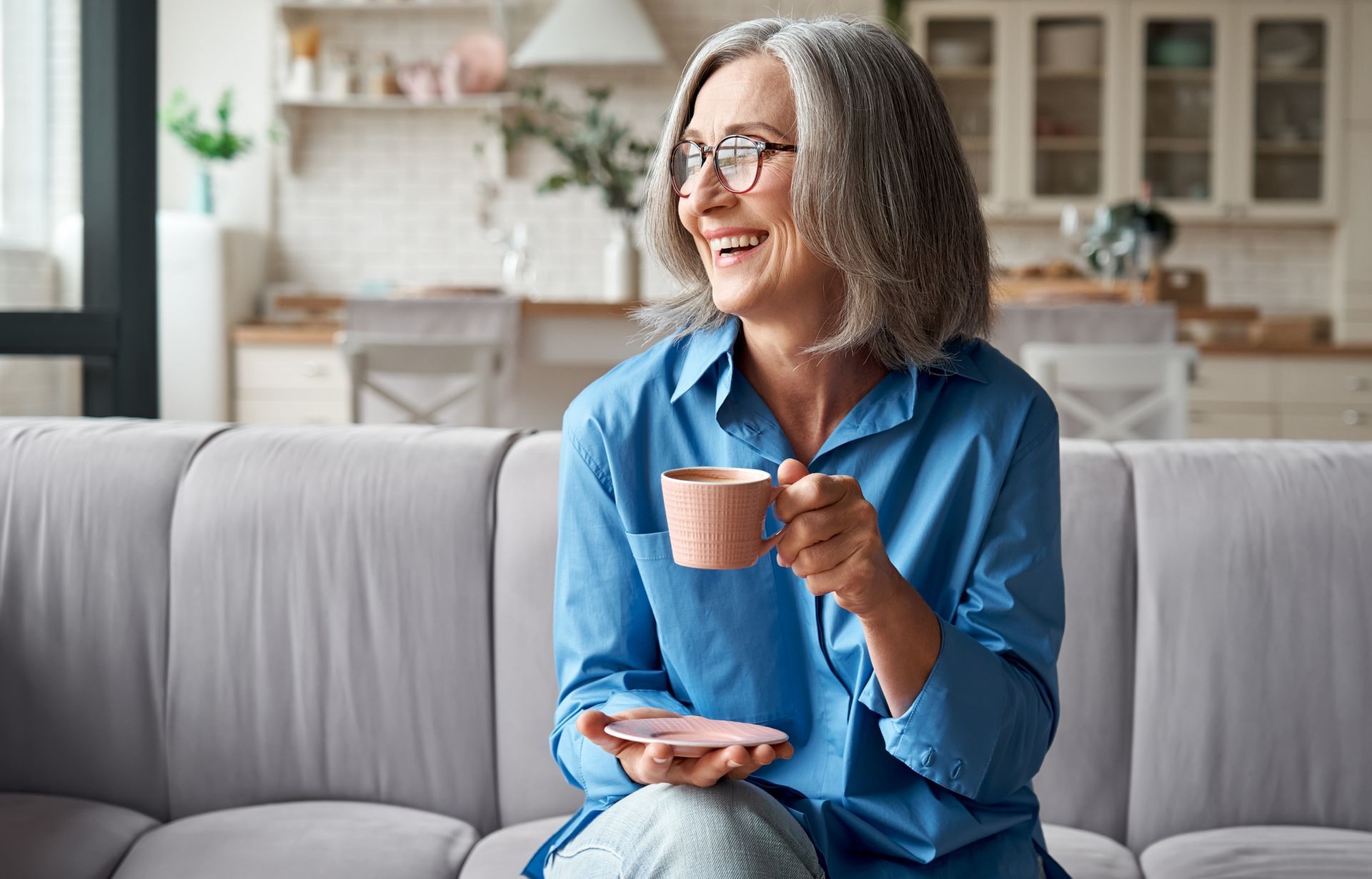 Woman with gray hair, holding a pink mug, laughing, sitting on a couch in a well-lit kitchen.
