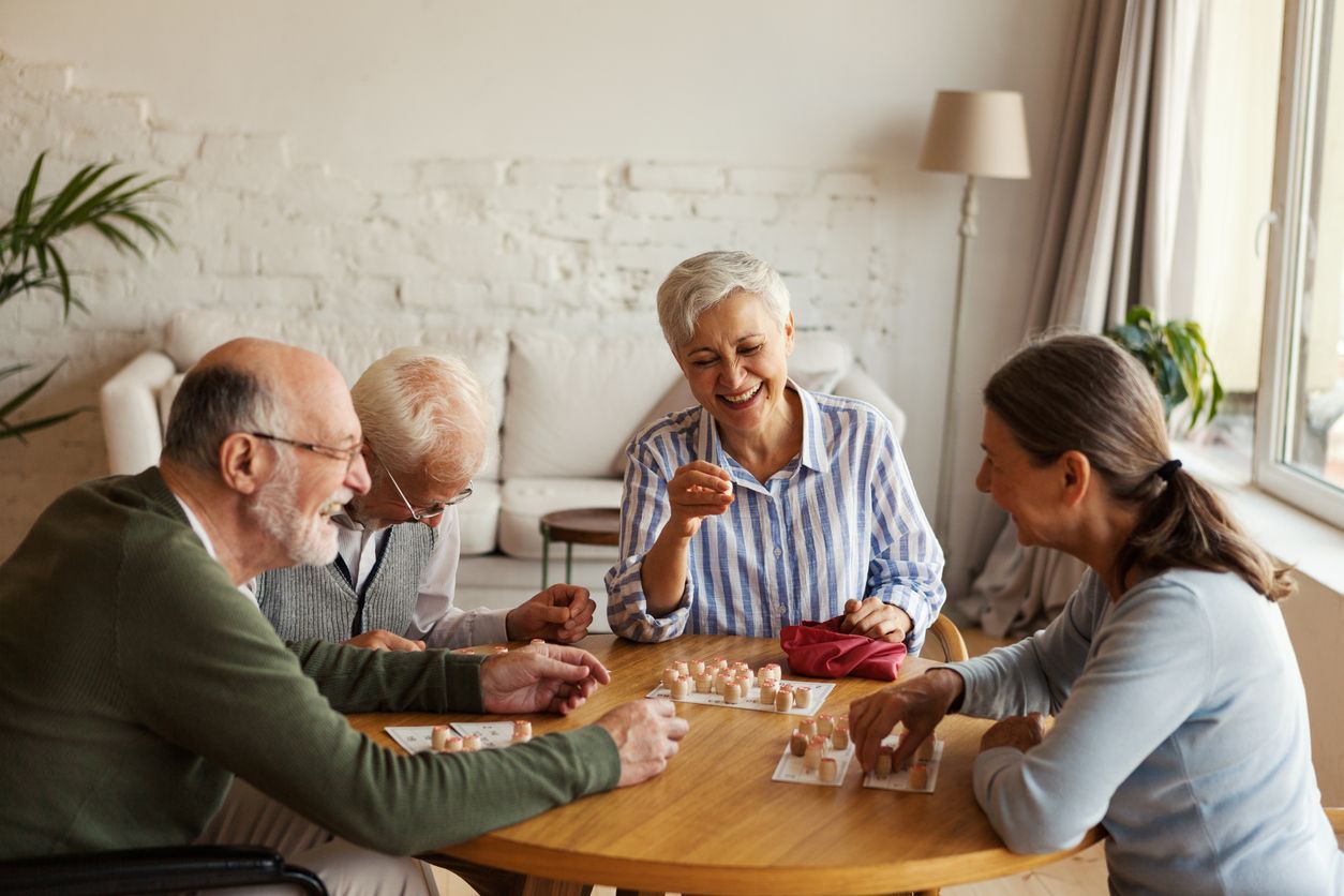 Four older adults laughing while playing a game at a round table. Bright, indoor setting.