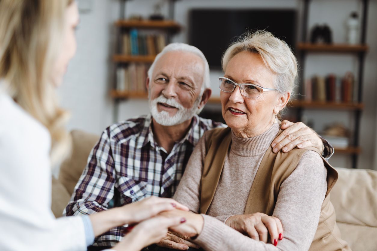 Senior couple consults with a healthcare provider, indoor setting. Man's arm around woman's shoulders; all are smiling.