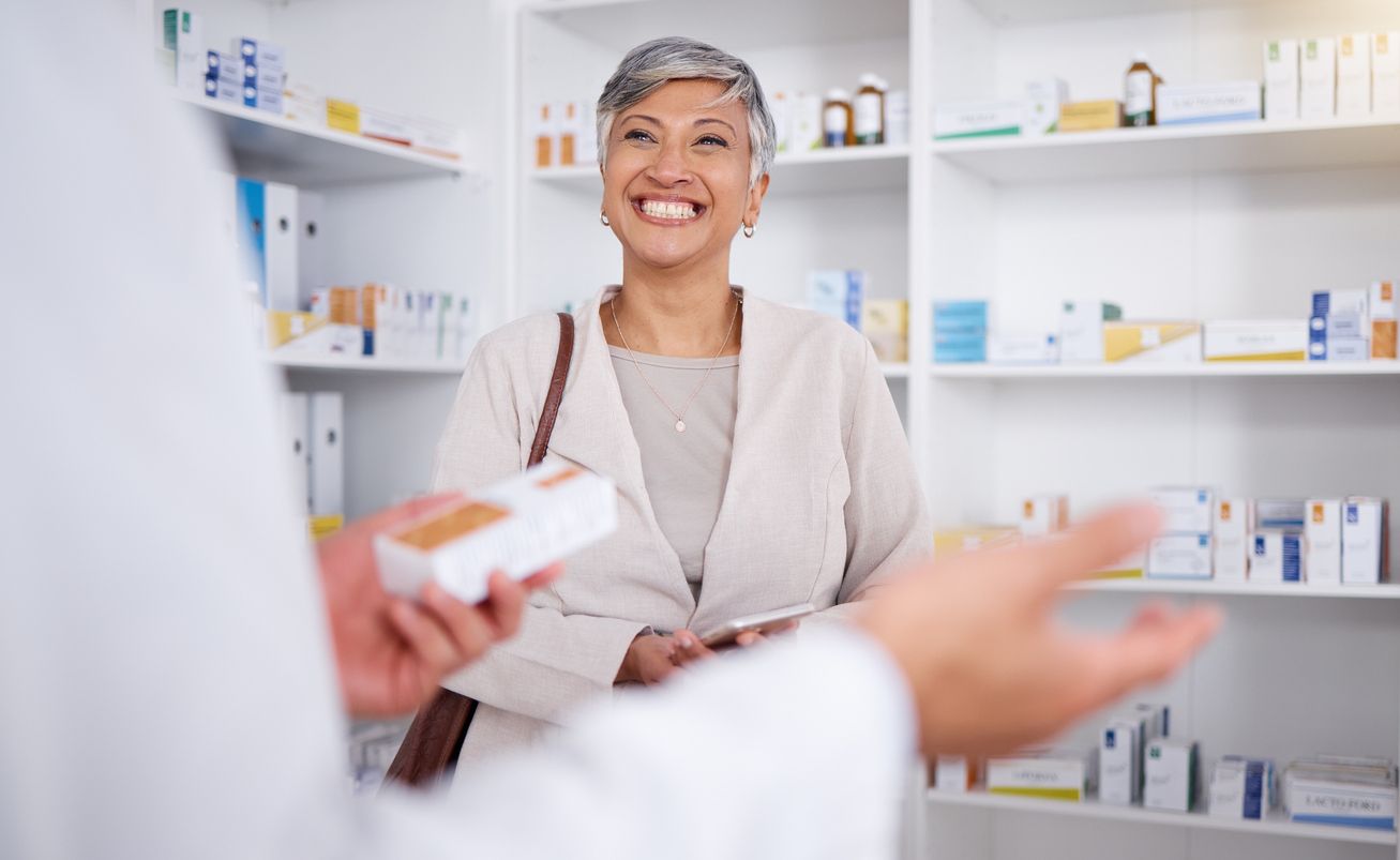A smiling woman at a pharmacy counter receives medication from a pharmacist. Shelves of products are in the background.