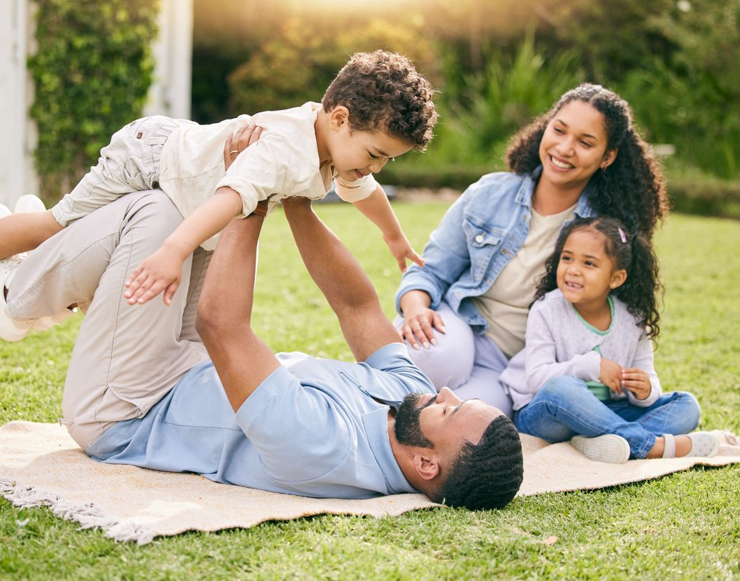 Father and son playing with a toy airplane in a sunny park. The boy raises the plane, smiling, while the father watches.