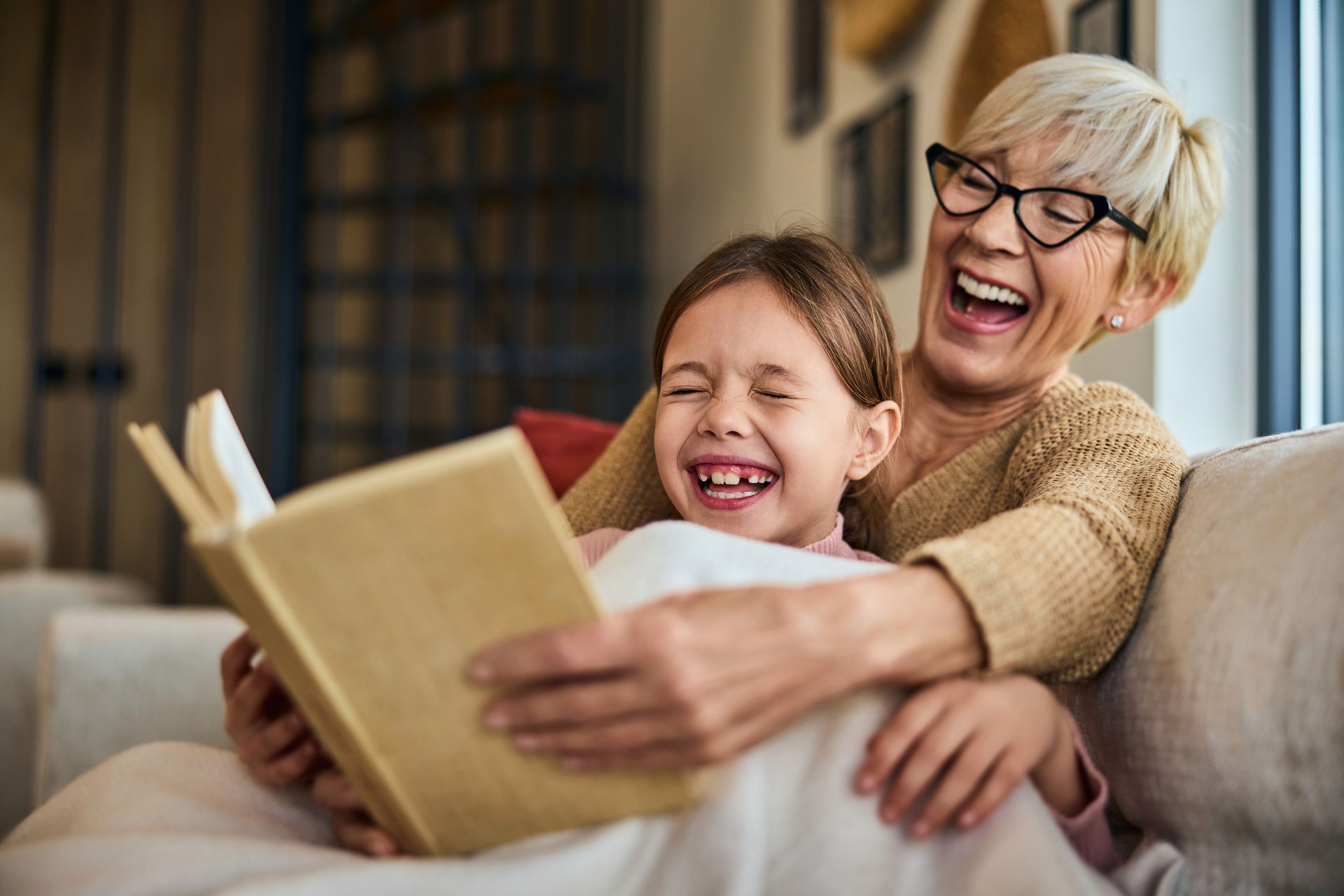 Grandmother and granddaughter laugh while reading a book together on a couch.