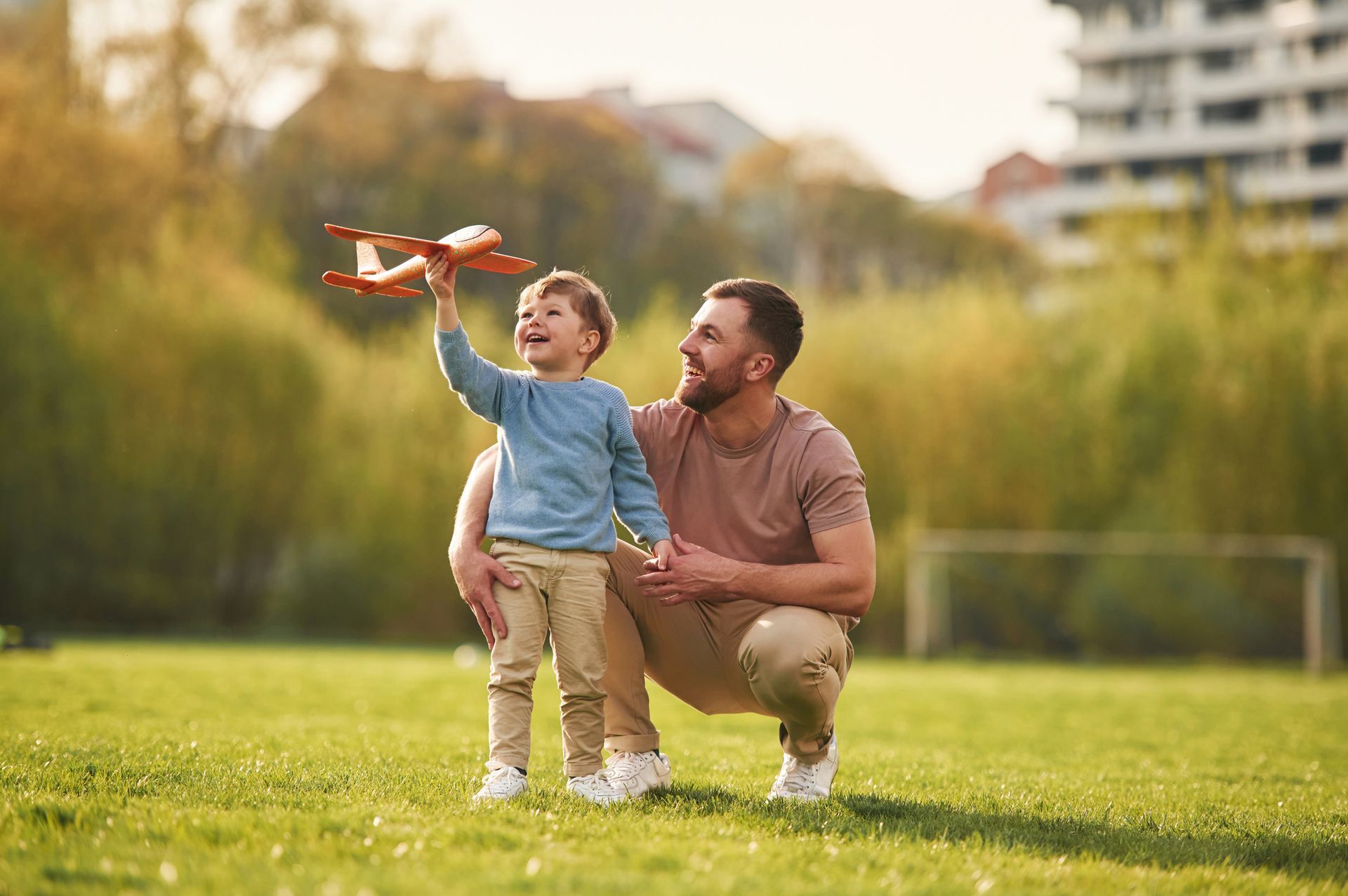Father and son playing with a toy airplane in a sunny park. The boy raises the plane, smiling, while the father watches.