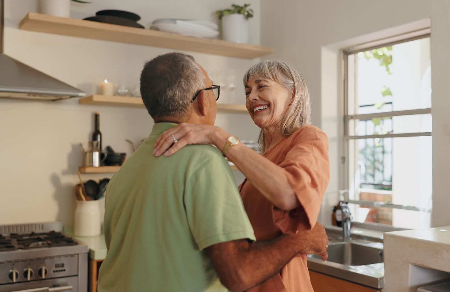 Senior couple dancing in kitchen, woman smiling, arms around each other, bright daylight.
