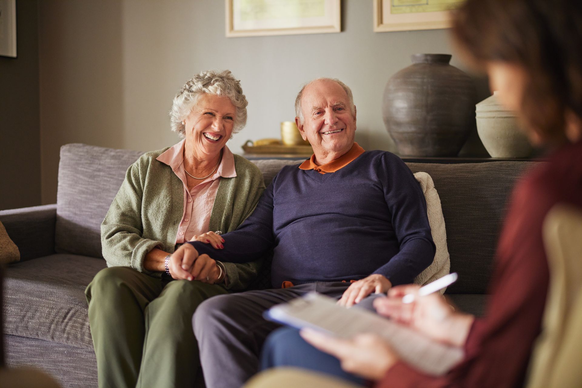 Elderly couple smiles while holding hands; seated on couch during a consultation.
