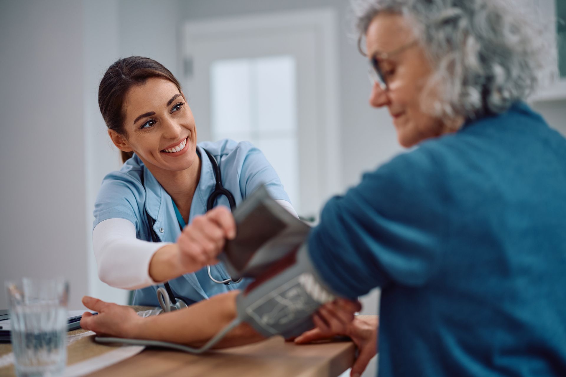 Nurse taking an elderly woman's blood pressure. Smiling nurse in blue scrubs, woman in blue, inside setting.