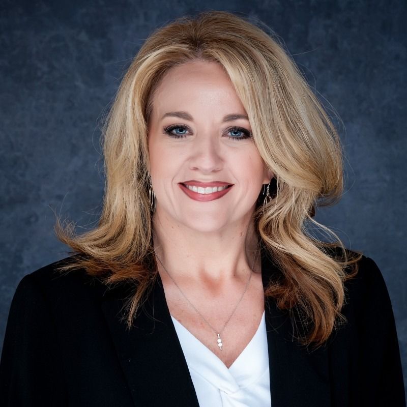 Blonde woman smiling, wearing a black blazer over a white top, against a blurred background.