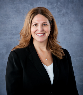 Woman with brown hair in a black suit smiling at the camera, against a blue-gray background.