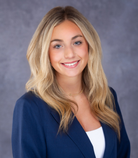Blonde woman in navy blazer smiles at camera; chest-up shot against gray backdrop.