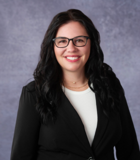 Woman with glasses, dark hair, wearing a black blazer and smiling in front of a blurred blue background.