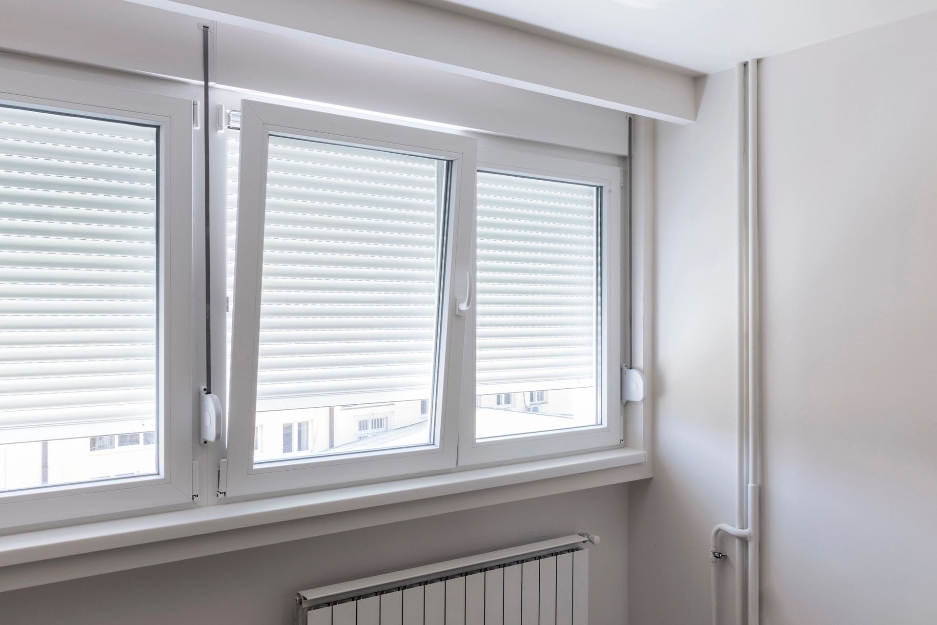 Finestra bianca con persiane chiuse, termosifone sotto, contro una parete grigio chiaro.