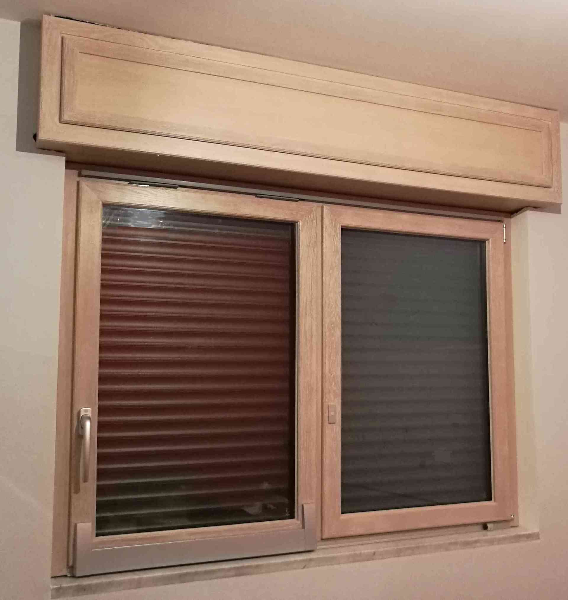 Finestra in legno con persiane chiuse, telaio di colore chiaro e alloggiamento superiore coordinato su una parete di colore chiaro.