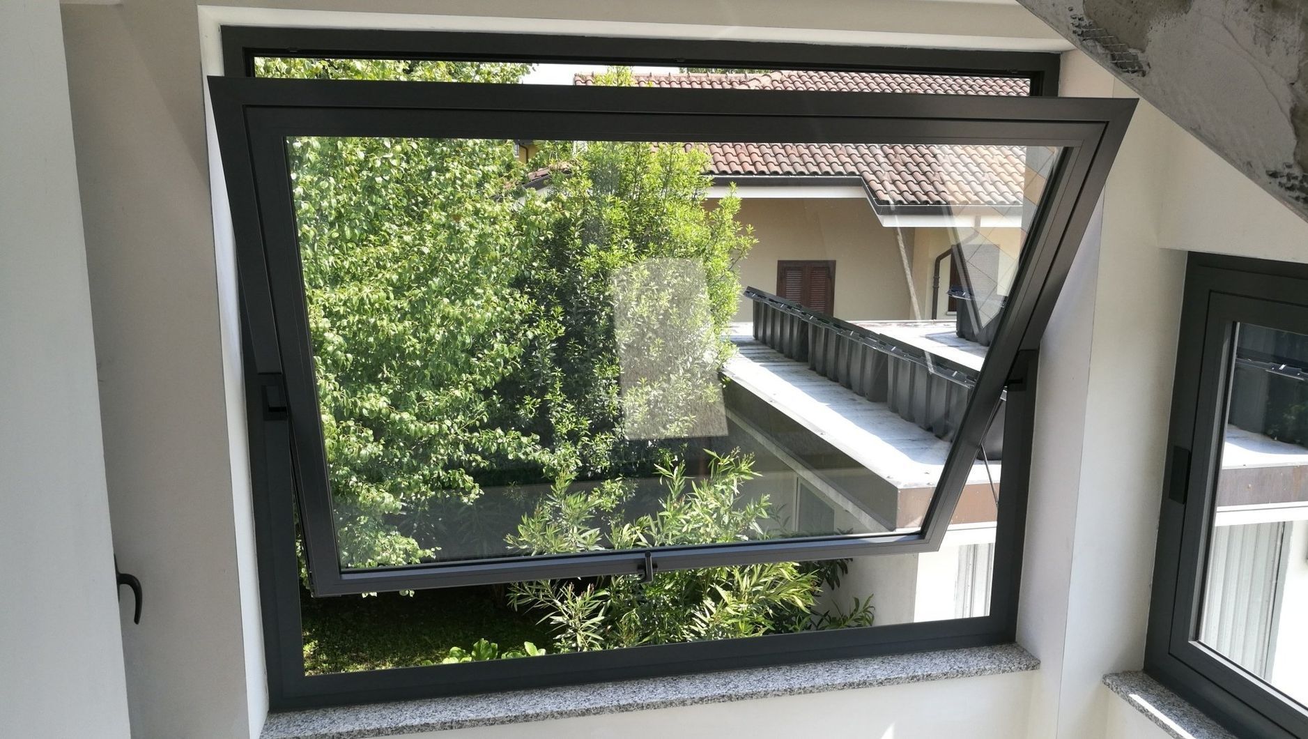 Una finestra aperta con cornice nera che si affaccia su alberi verdi e sul balcone di un edificio.