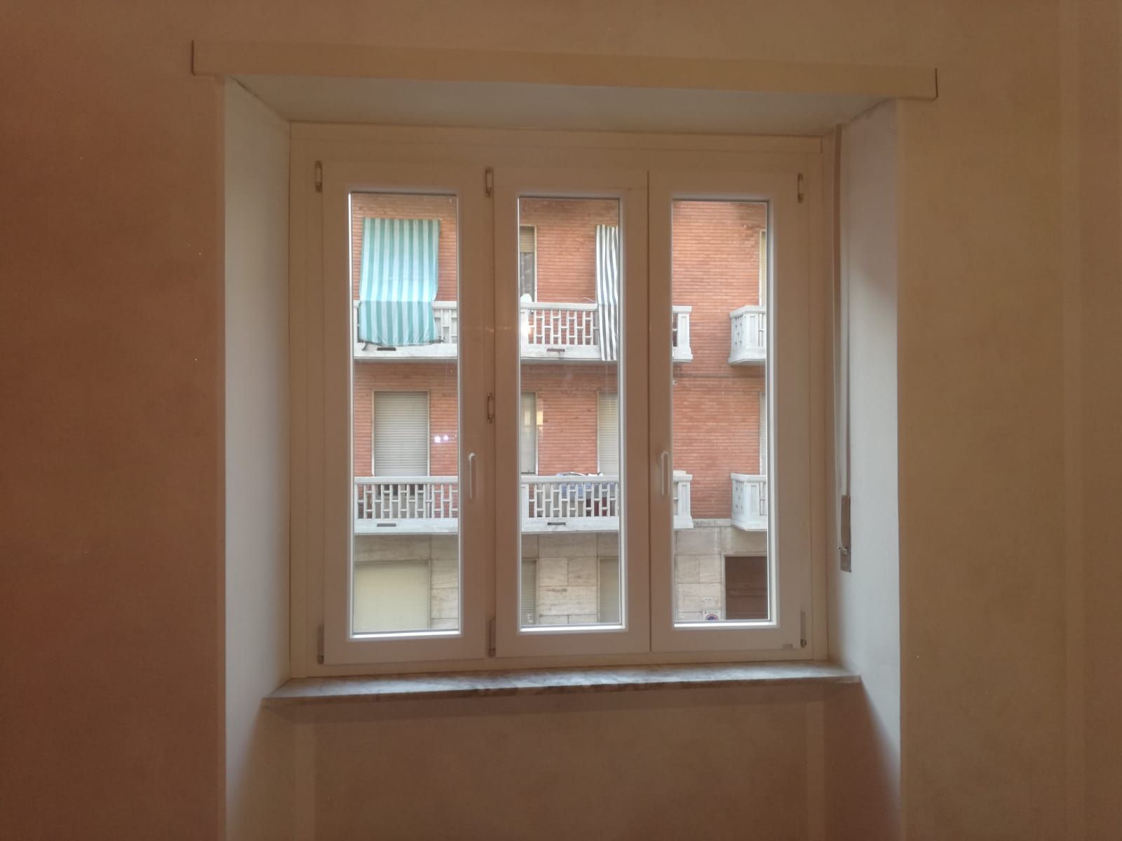 Finestra a triplo vetro con persiane grigie chiuse; telaio bianco. Vista sul verde esterno attraverso la finestra.