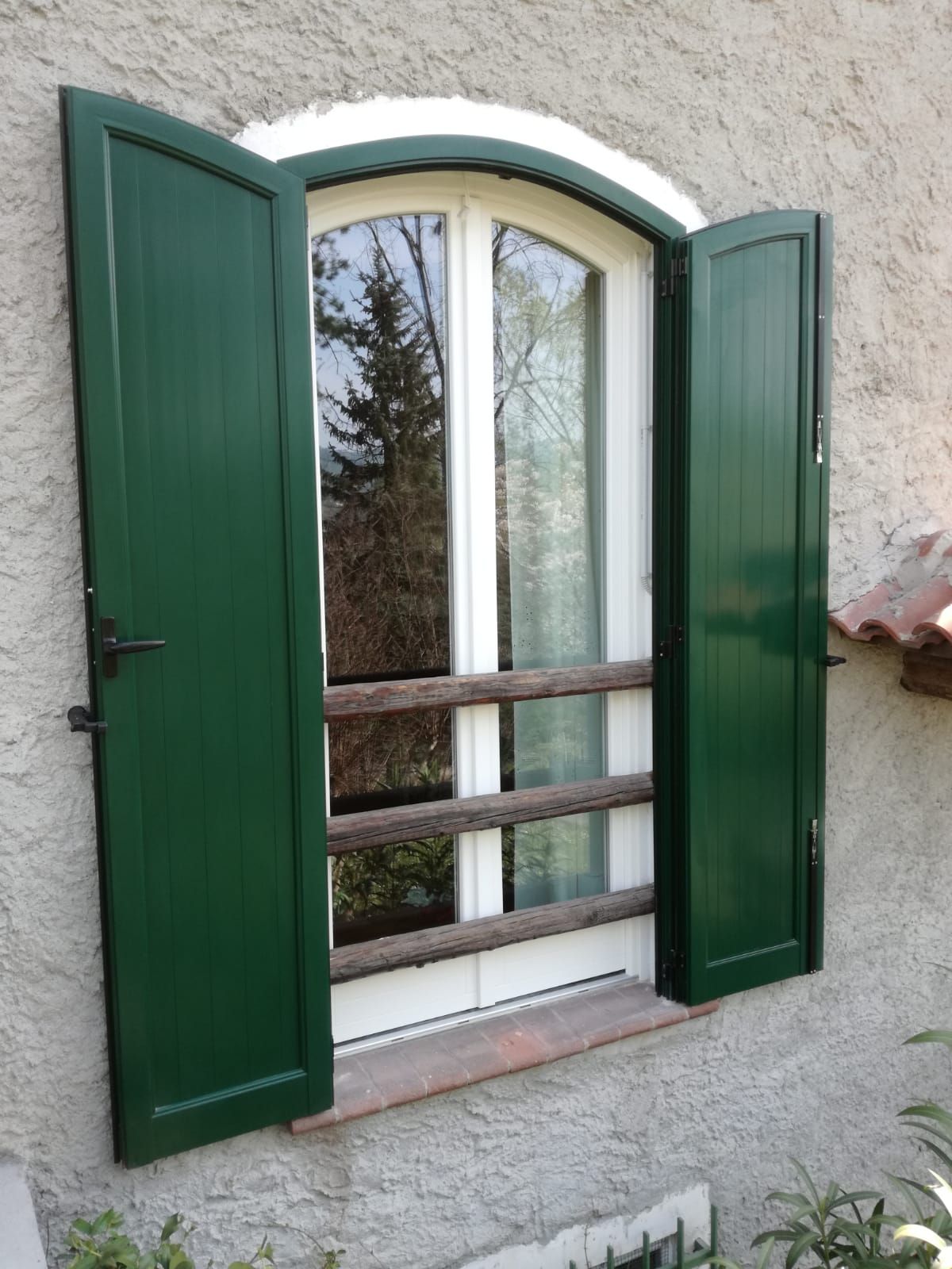 Persiane verdi si aprono su una finestra bianca ad arco. Un albero si riflette nel vetro.