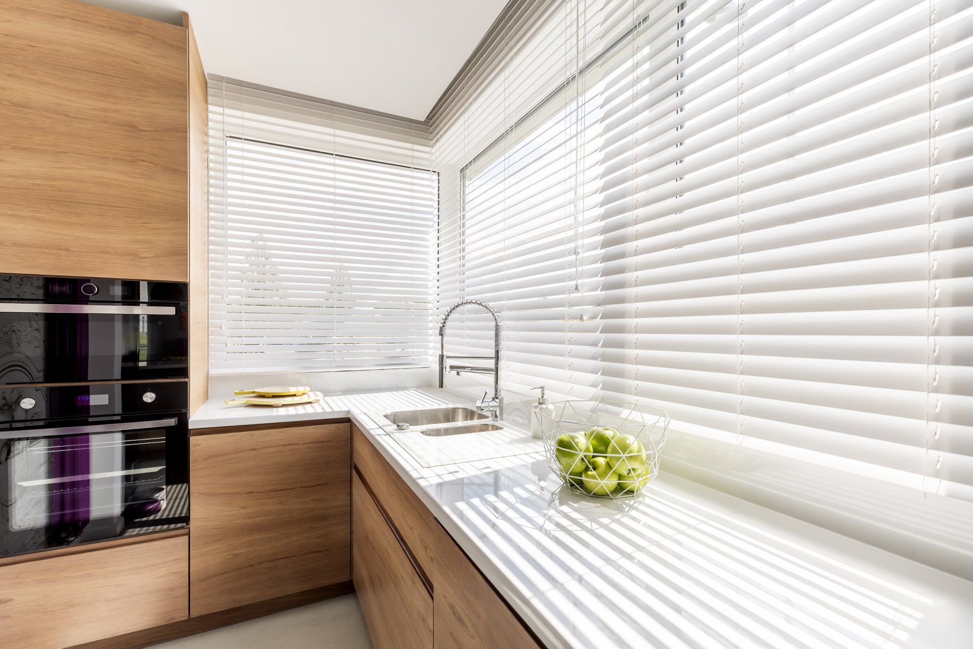 Angolo cottura con tende bianche, mobili, lavello e piano di lavoro. Una mela è appoggiata sul piano di lavoro.