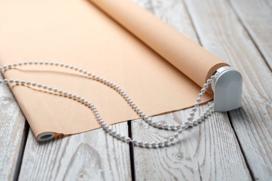 Tenda a rullo beige parzialmente srotolata su una superficie di legno, con cordino di trazione decorato con perline.