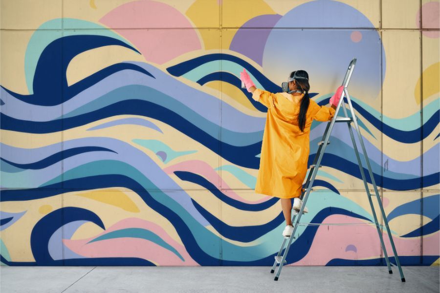 A woman is standing on a ladder painting a mural on a wall.