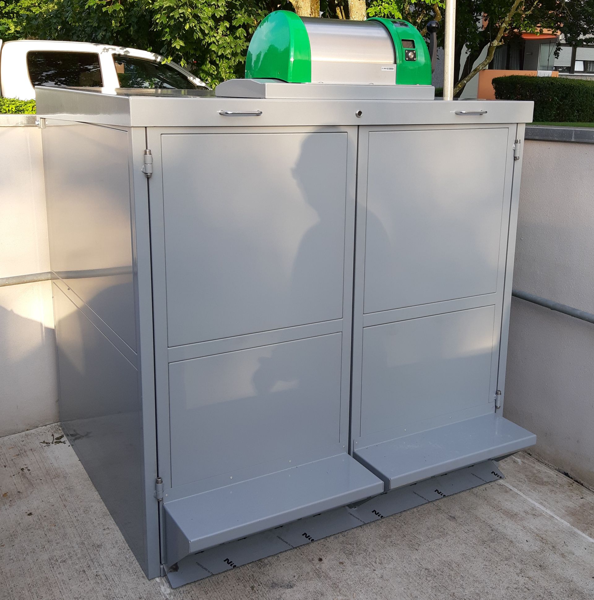 Enceinte grise pour conteneurs de recyclage avec deux portes, pédales et couvercle vert, installée en extérieur.