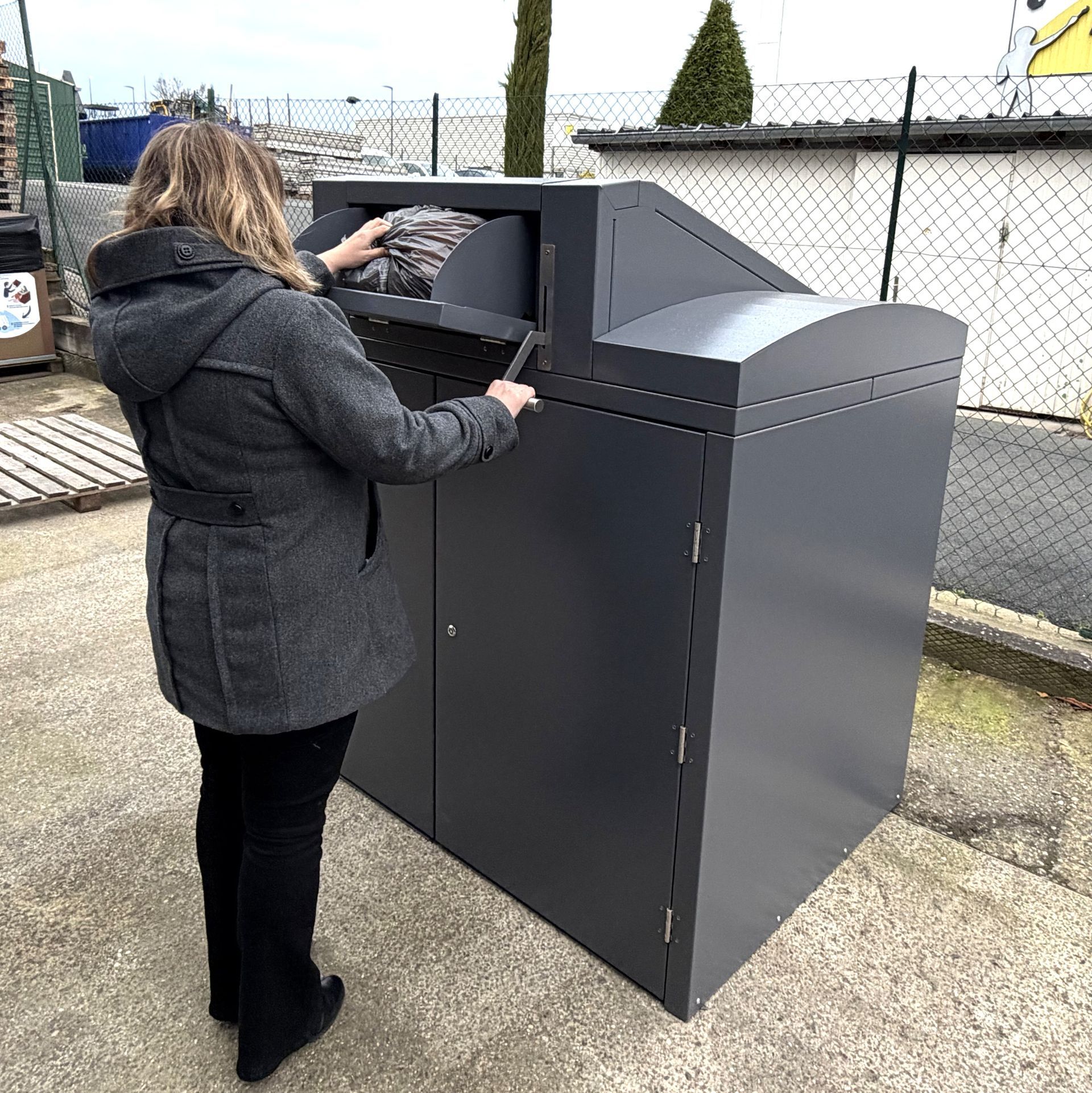 Une femme dépose des objets dans un bac de recyclage gris foncé à l'extérieur.