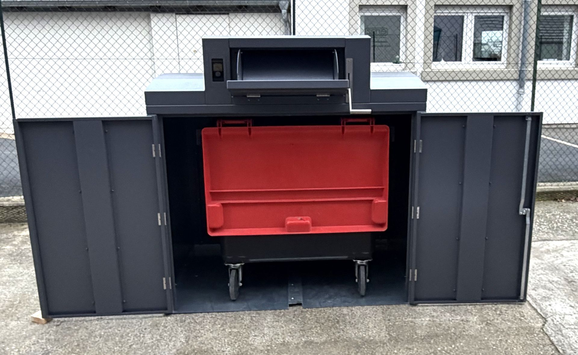 Un conteneur à déchets gris foncé, portes ouvertes laissant apparaître une poubelle rouge à l'intérieur, placé à l'extérieur près d'une clôture.