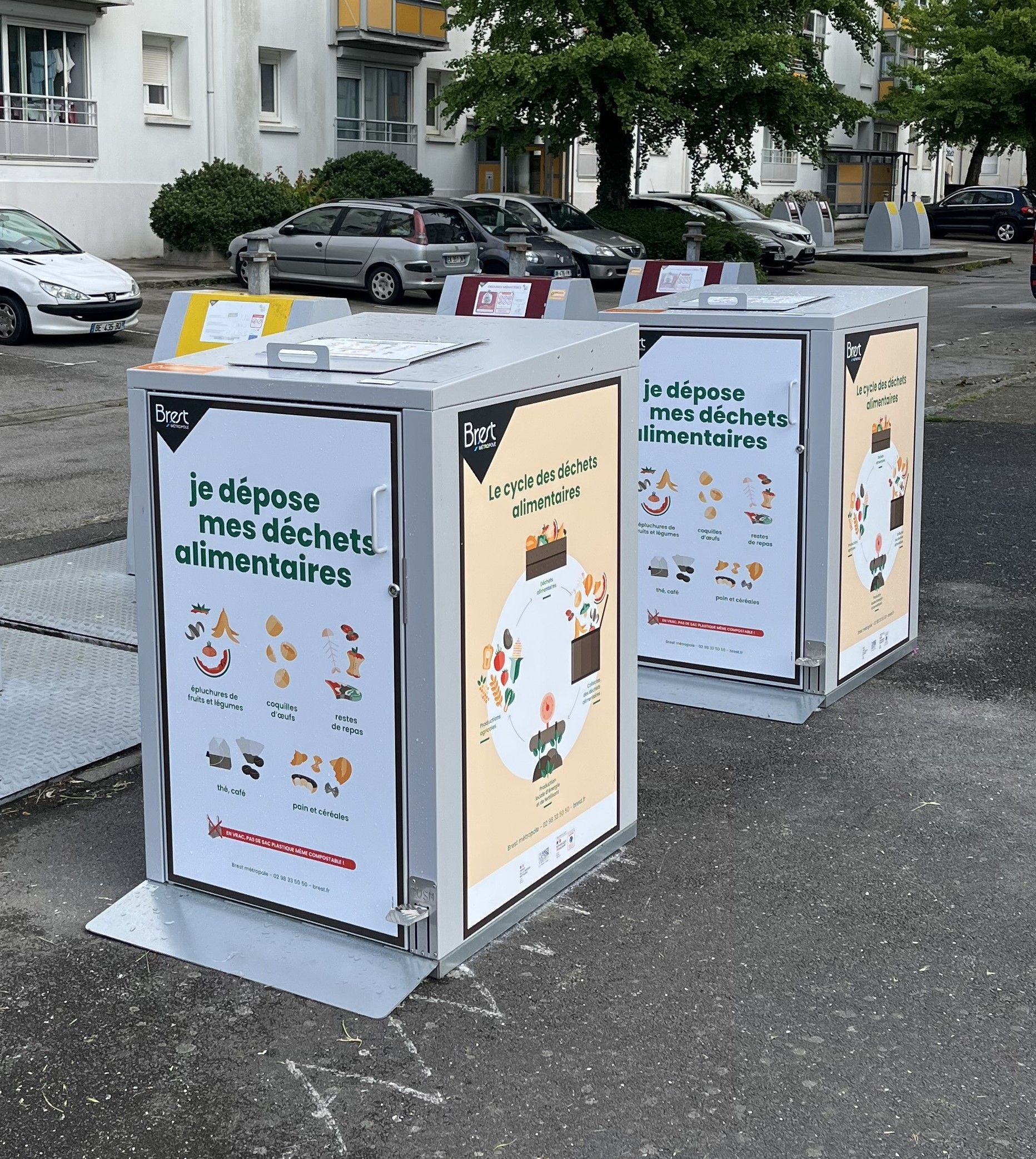 Deux conteneurs de recyclage en métal avec des affiches informatives dans un espace extérieur ; des voitures garées en arrière-plan.
