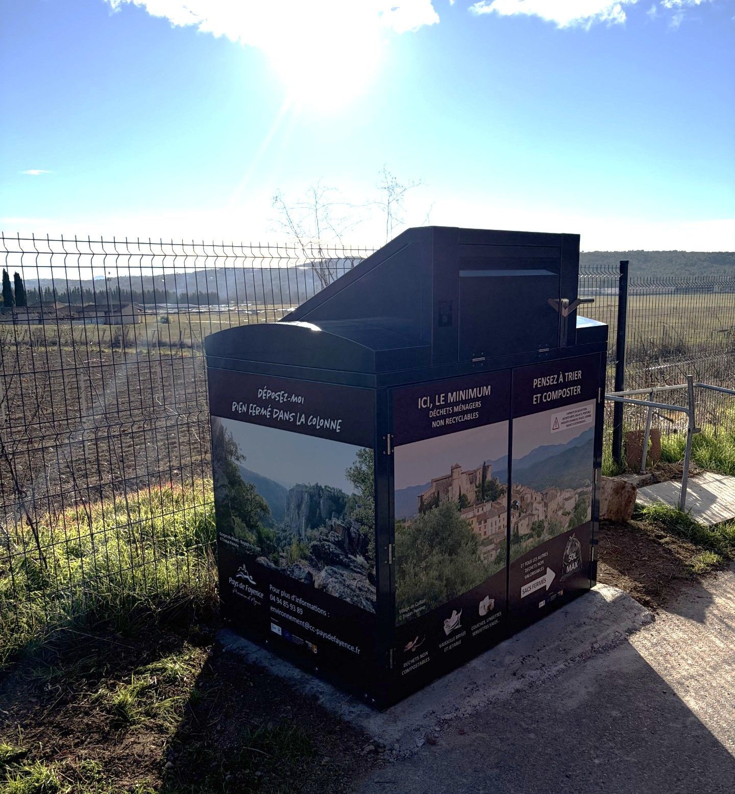 Kiosque d'information noir orné de photos de paysages, situé à côté d'un vignoble, sous un soleil éclatant.