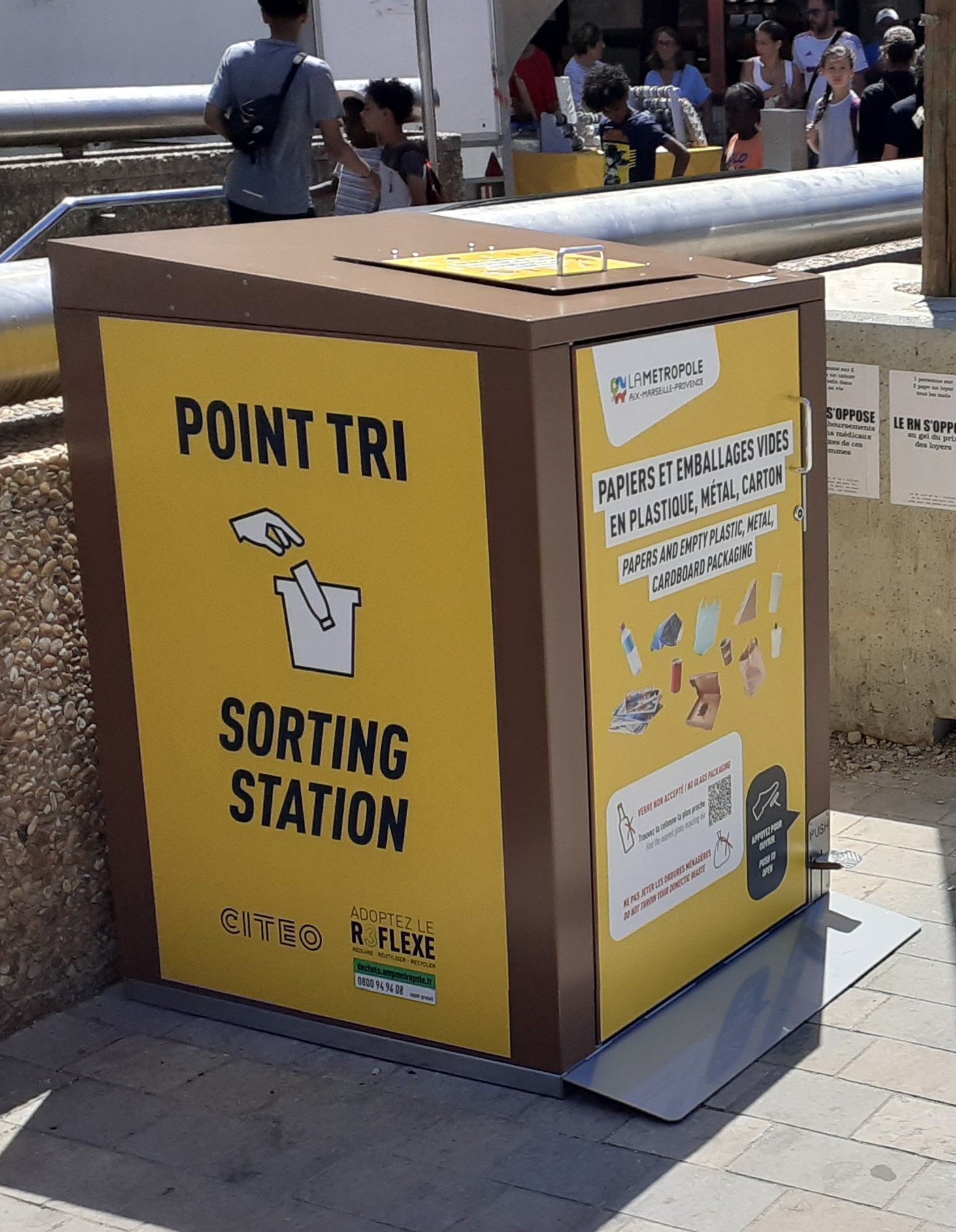 Station de tri des déchets recyclables marron et jaune dans un espace public extérieur.