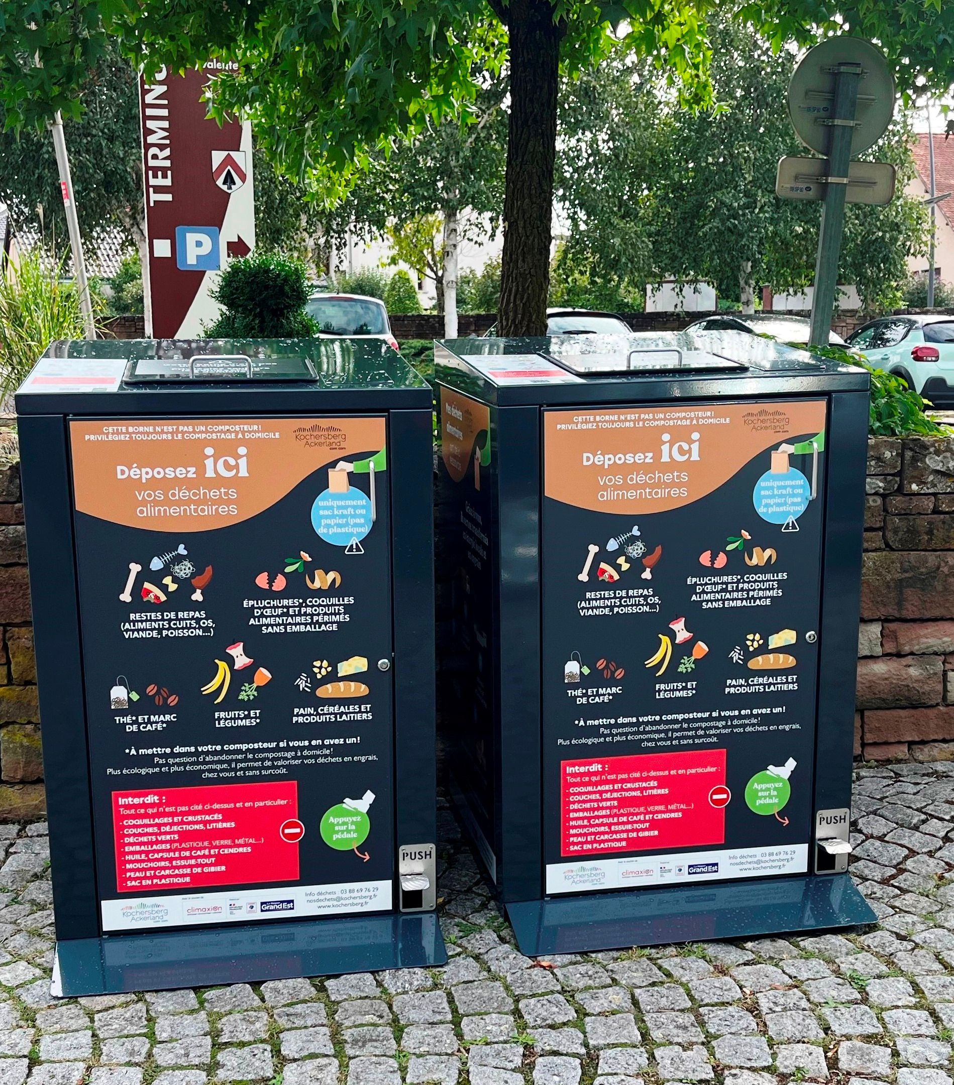 Deux grandes poubelles de recyclage noires avec des panneaux d'instructions sur une rue pavée.