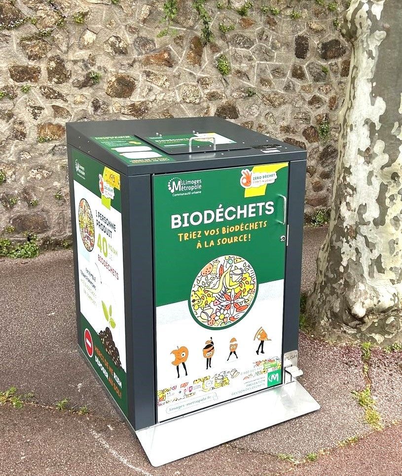 Poubelle verte de recyclage des « biodéchets » (déchets alimentaires) sur un trottoir devant un mur en pierre.