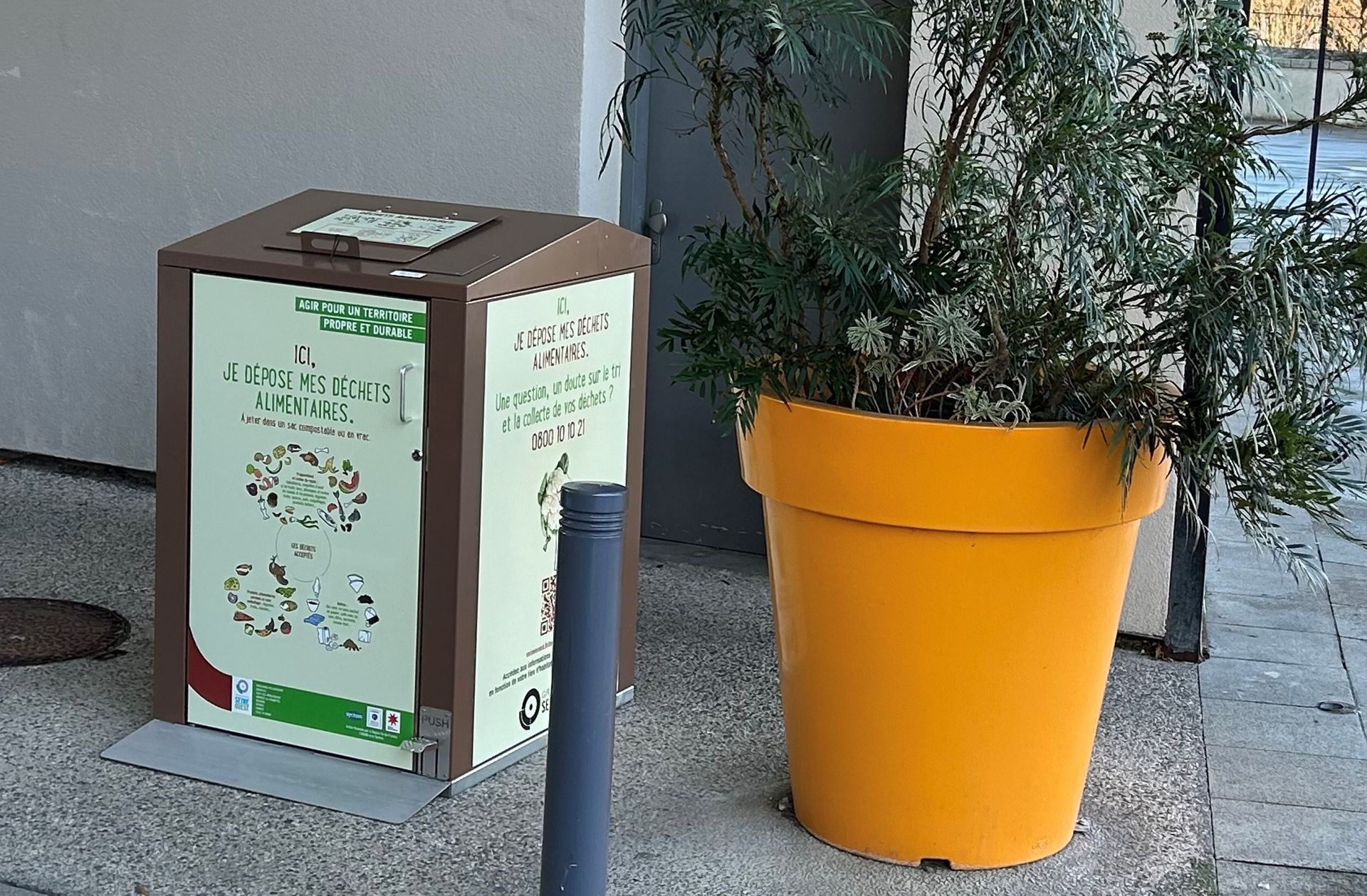 Quatre poubelles de recyclage vertes à couvercles noirs sont disposées sur un trottoir, à côté d'un bâtiment en briques.
