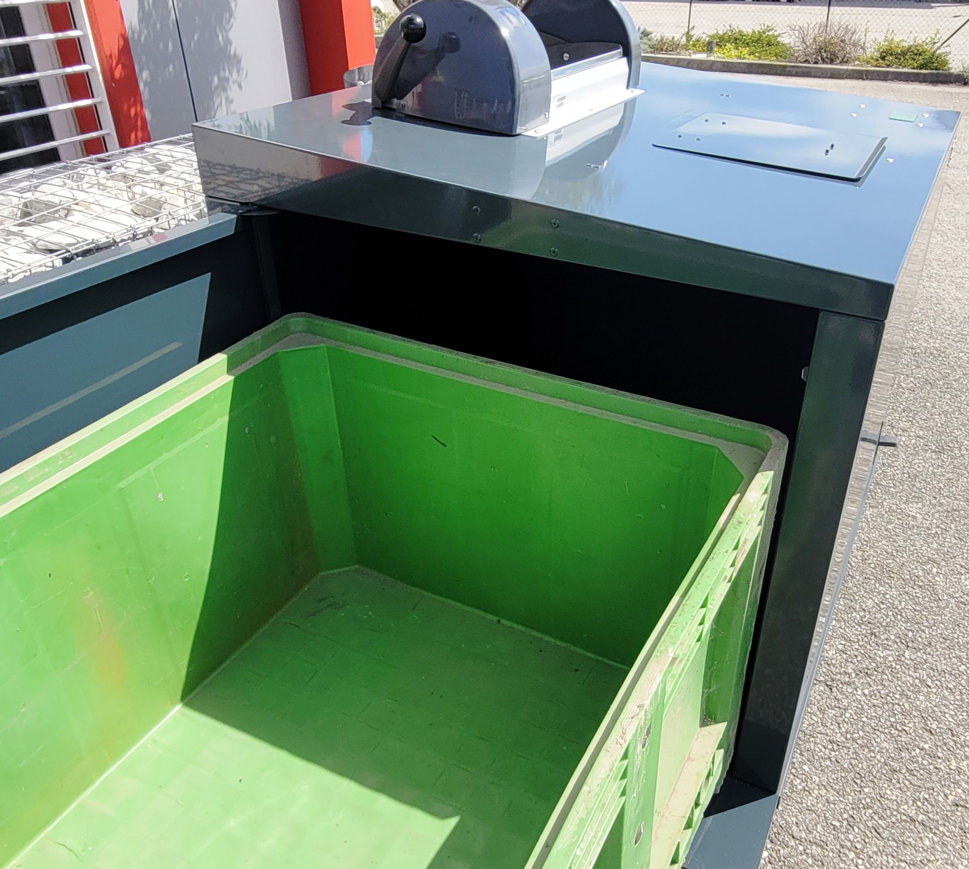 Poubelle de recyclage verte à l'intérieur d'un boîtier métallique foncé, couvercle et poignée visibles.