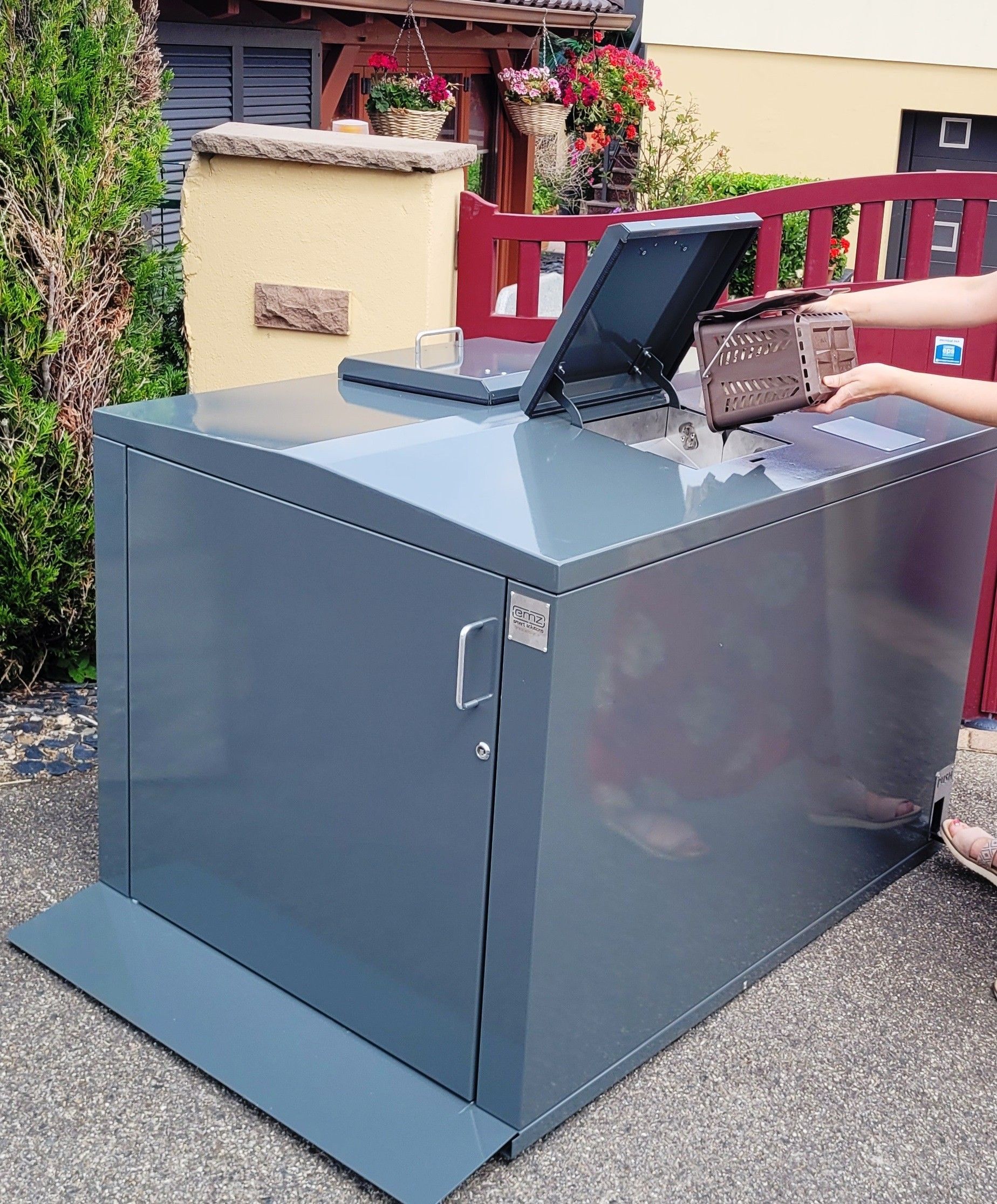 Boîte de dépôt de colis en métal gris avec couvercle et porte ouverts, une personne tend la main à l'intérieur.