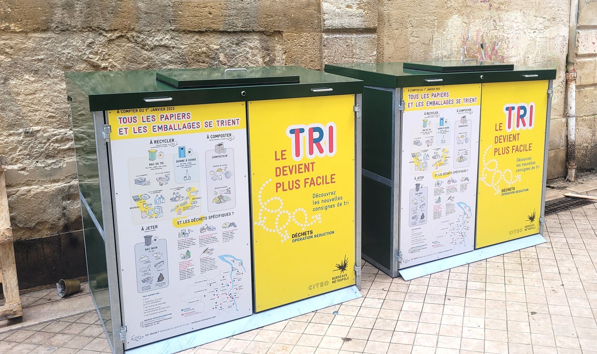 Bacs de recyclage, panneaux jaunes et blancs. Étiquette « TRI », illustrations des articles recyclables. À l'extérieur, près d'un mur en pierre.