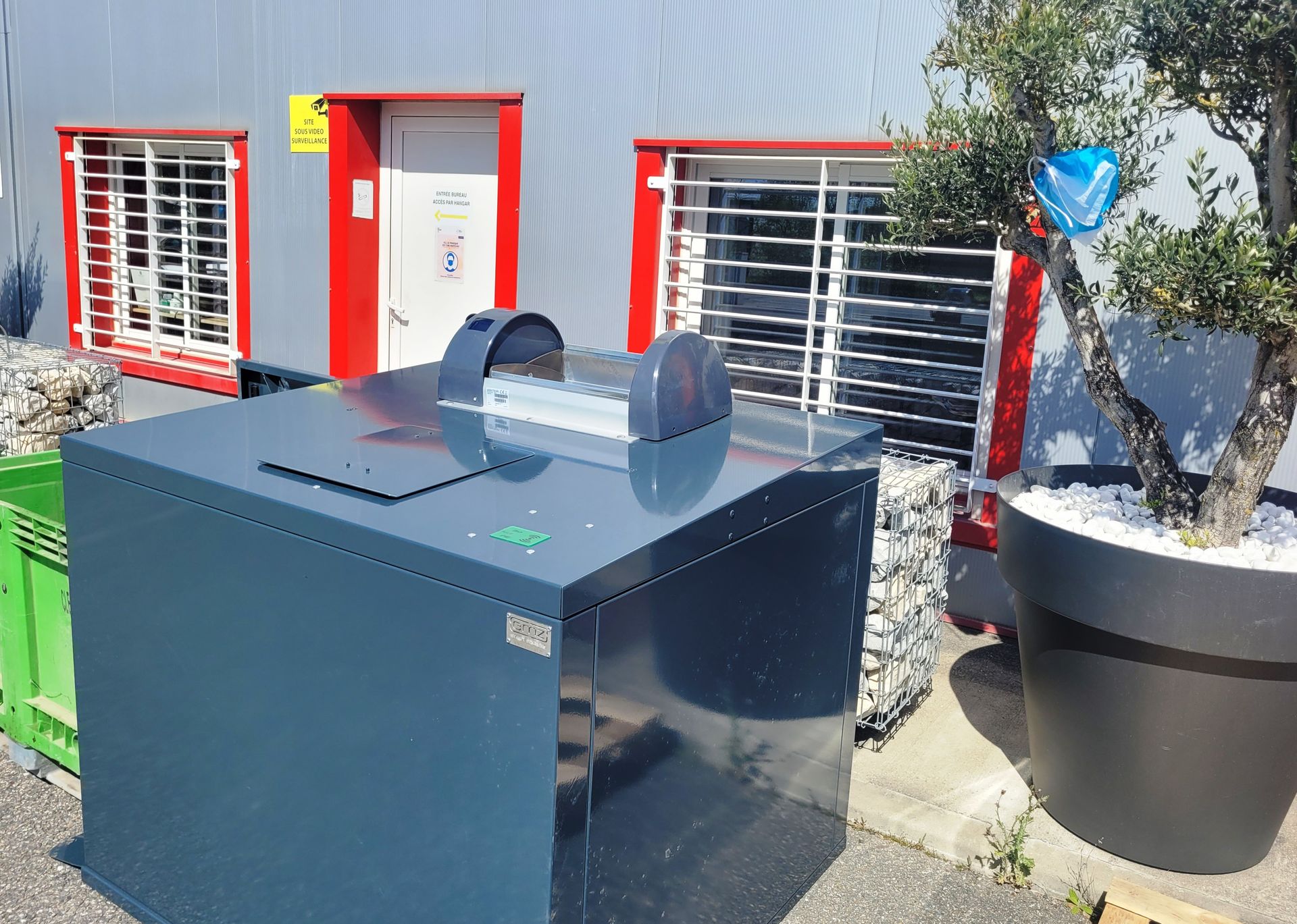 Une déchiqueteuse à papier grise à l'extérieur d'un bâtiment avec des barreaux de sécurité et un arbre en pot.