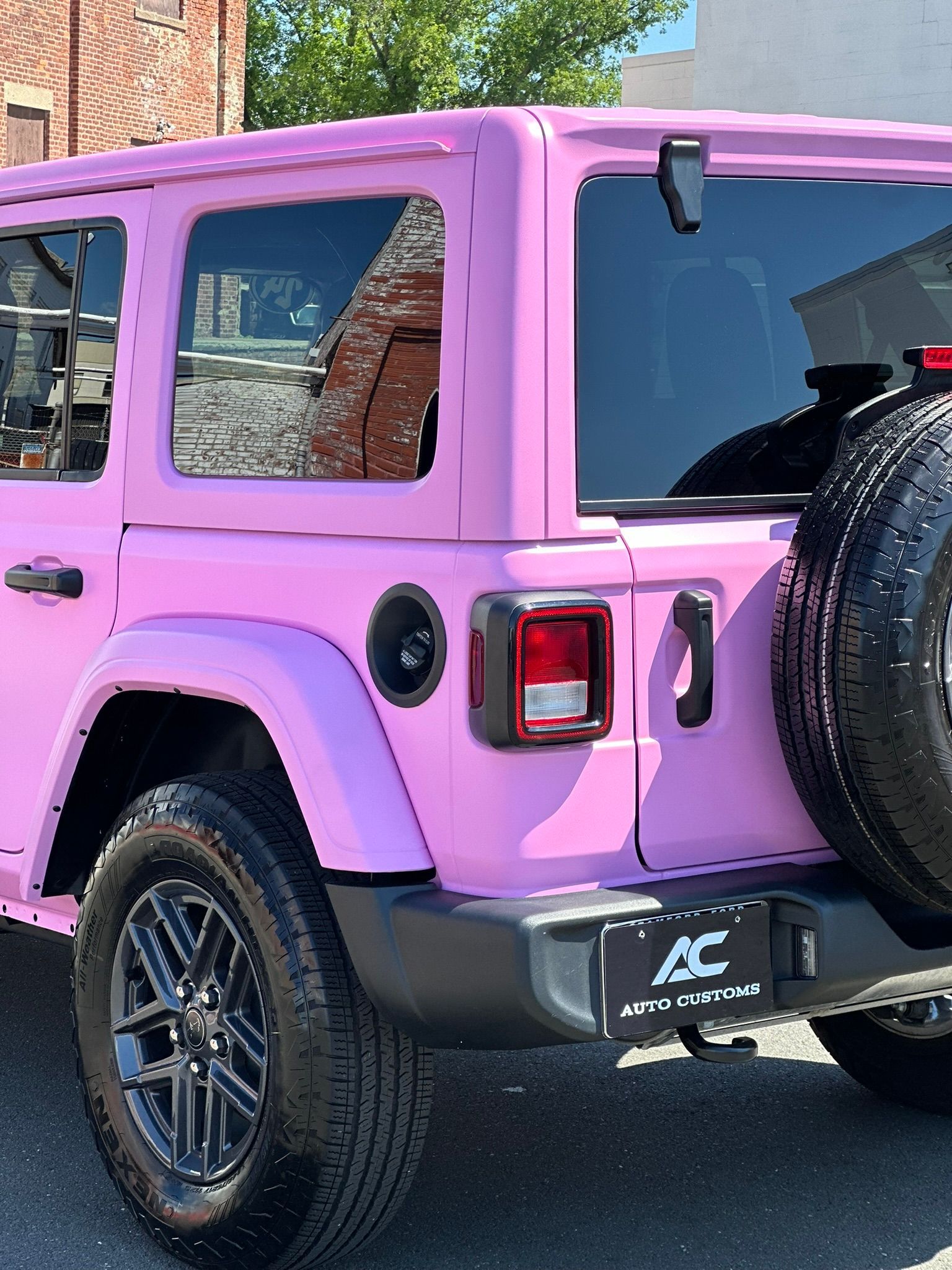 Pink Jeep Wrangler parked on a street, black accents, spare tire, and a black bumper.