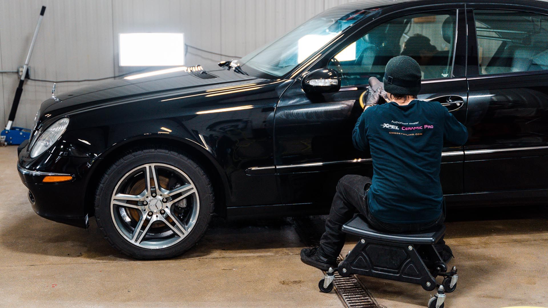 A man is sitting on a stool in front of a black car.