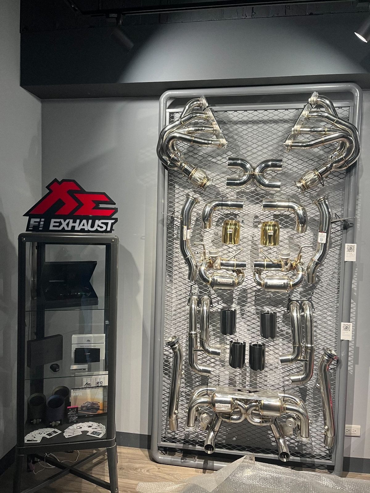 A shiny, complex car exhaust system displayed on a metal grid in a shop, next to a branded sign.