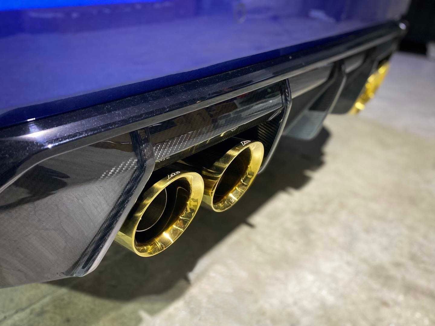 Close-up of a car's rear end with gold exhaust tips against a carbon fiber and blue body.