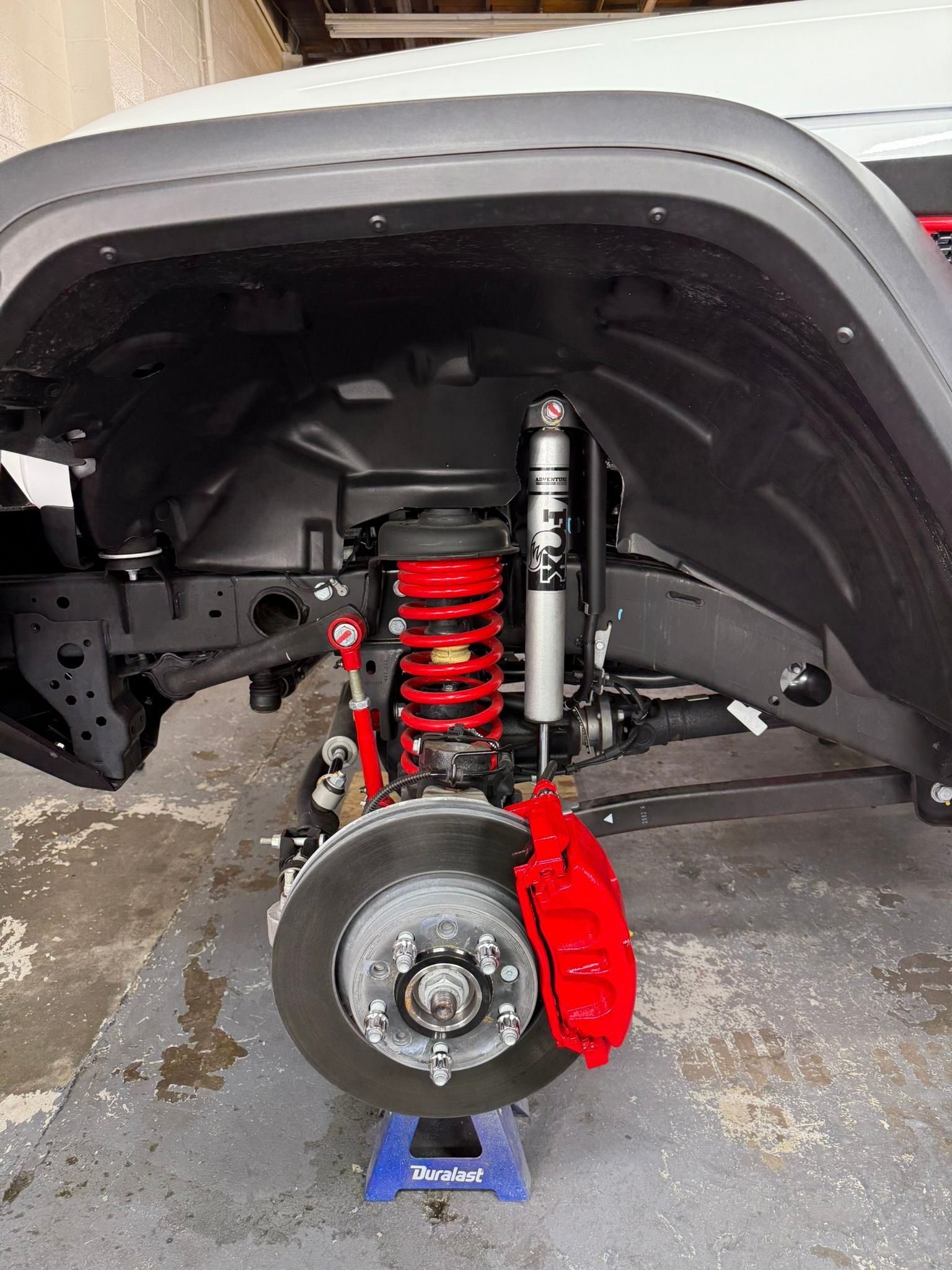 Red and black suspension components visible on a vehicle's undercarriage. The wheel and tire are removed.