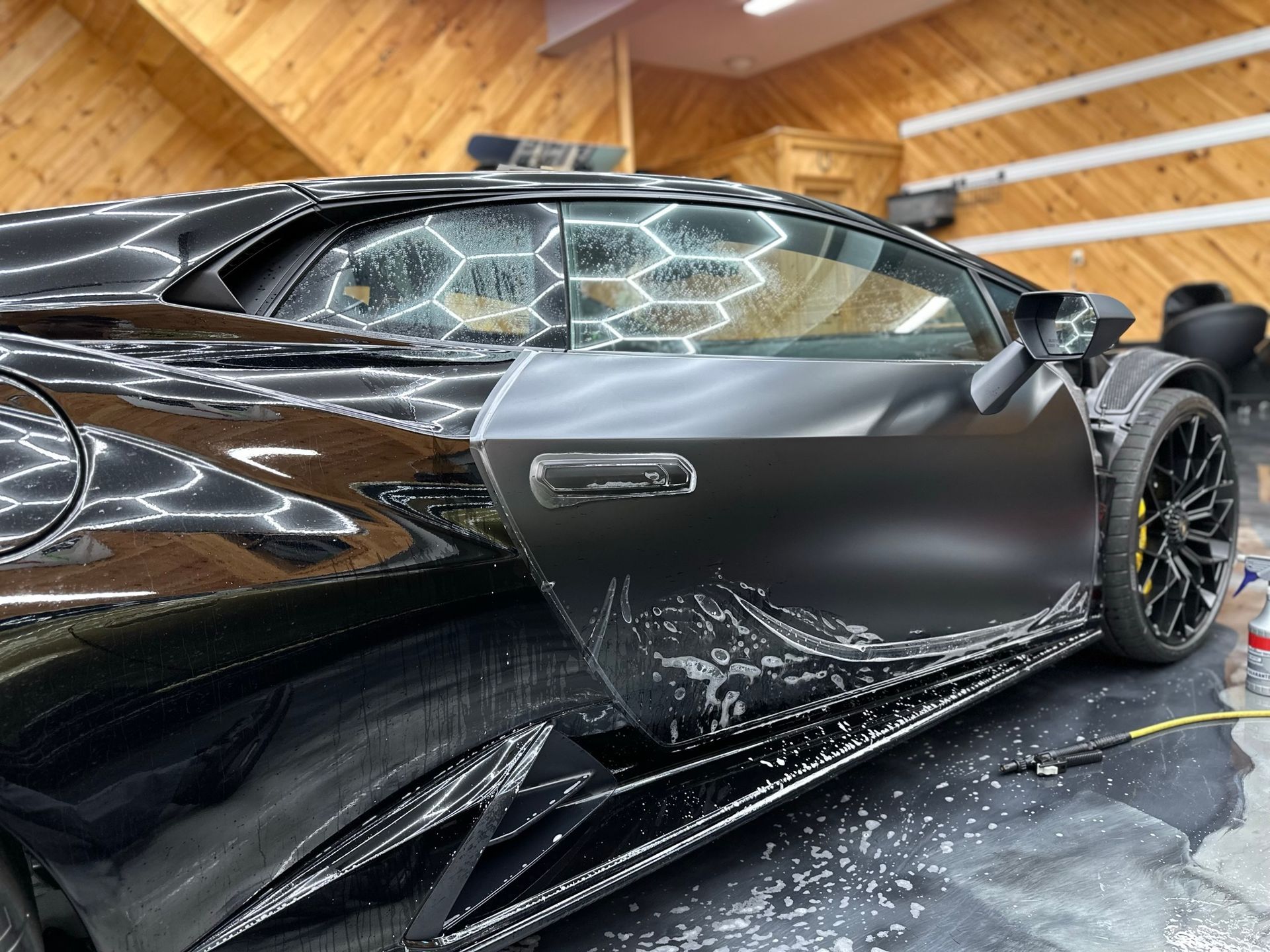 Black Lamborghini with a door open, undergoing paint protection film application in a workshop.